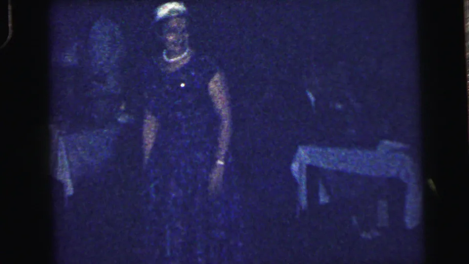 1964 Camden home-movie frame showing silhouetted adults at a dark reunion dance, with one woman and a table faintly visible.
