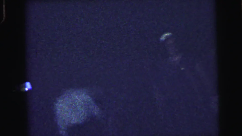 Low-light 8mm frame from the reunion dance, mostly shadowed with a small bright patch and minimal visible detail.