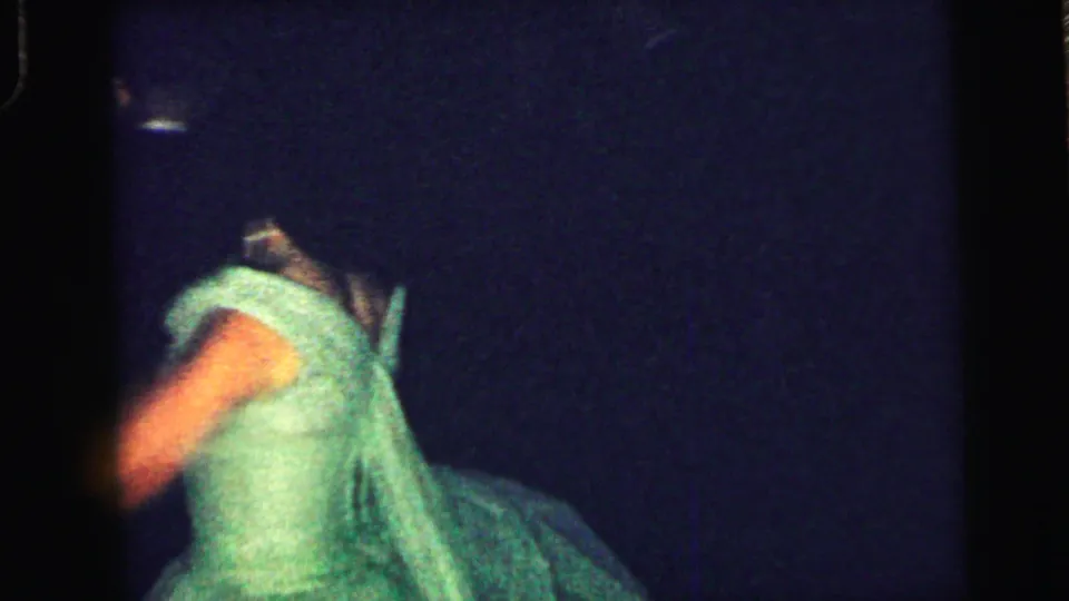 A woman in a green formal dress dances in a dim Camden, New Jersey party setting, recorded on 8mm film in 1964.