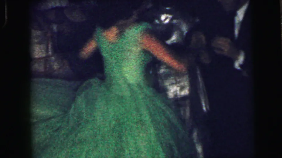 1964 home-movie frame from Camden showing a green-dressed dancer moving in front of suited guests indoors.