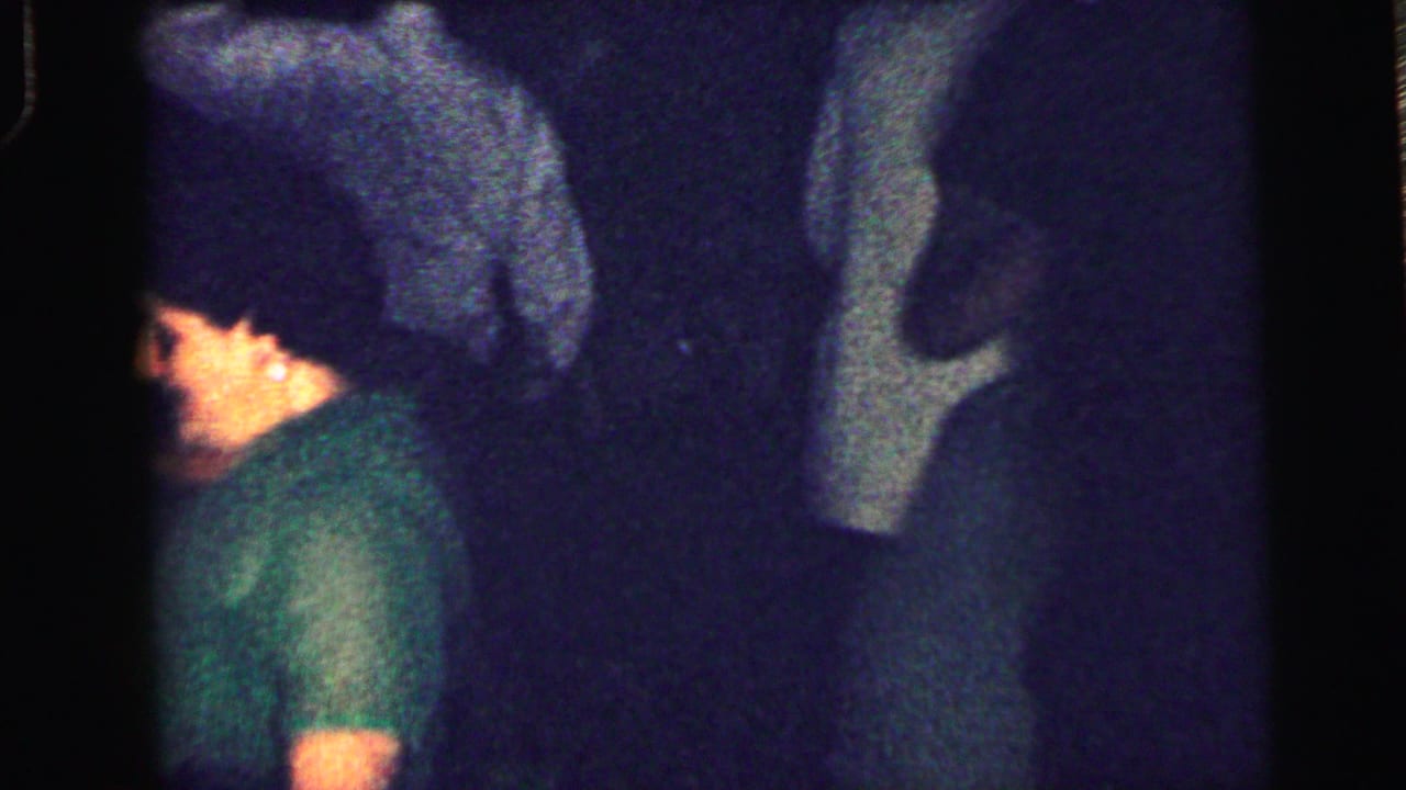 A young man in a green shirt is seen in profile among other partygoers in the low-light 1964 Camden footage.