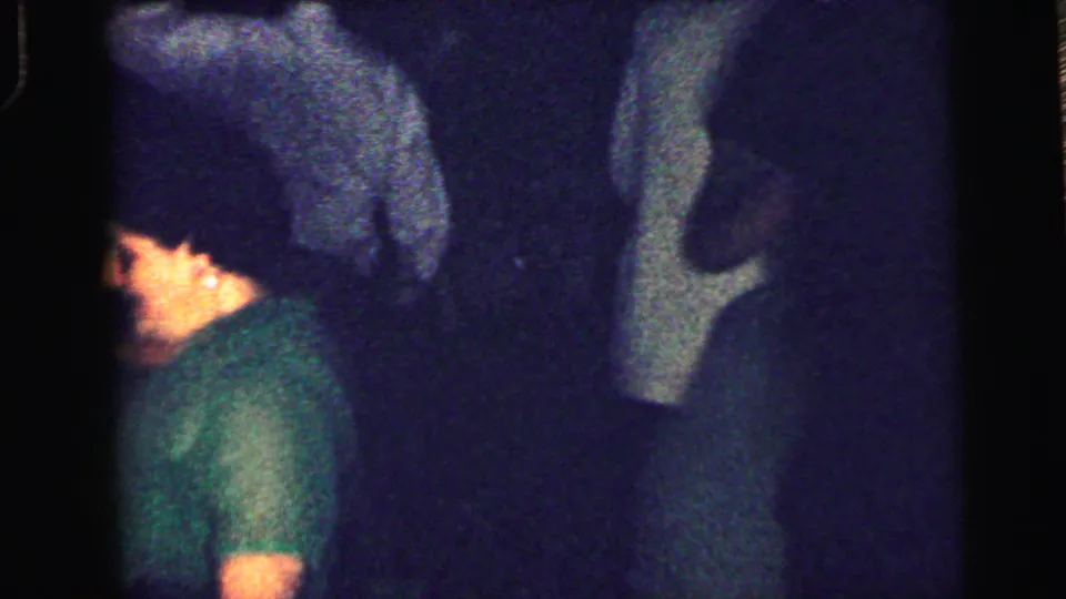 A young man in a green shirt is seen in profile among other partygoers in the low-light 1964 Camden footage.