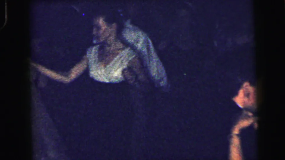 A woman in a light blouse gestures while dancing or socializing in the dim 1964 Camden home-movie party scene.