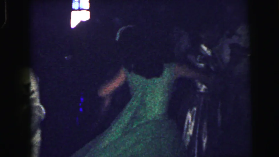 1964 Camden 8mm frame showing a woman in a green formal dress dancing near other guests under dim interior lighting.