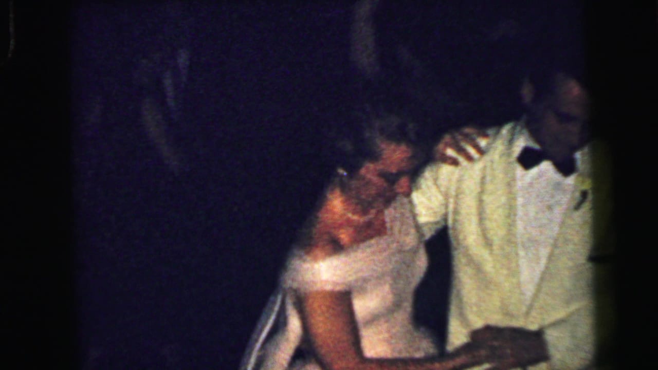 Woman in a white gown dances beside a man in a light tuxedo and bow tie.