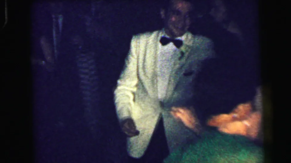 1964 Camden home-movie frame showing a man in a light tuxedo and bow tie near a dance partner in dim party light.