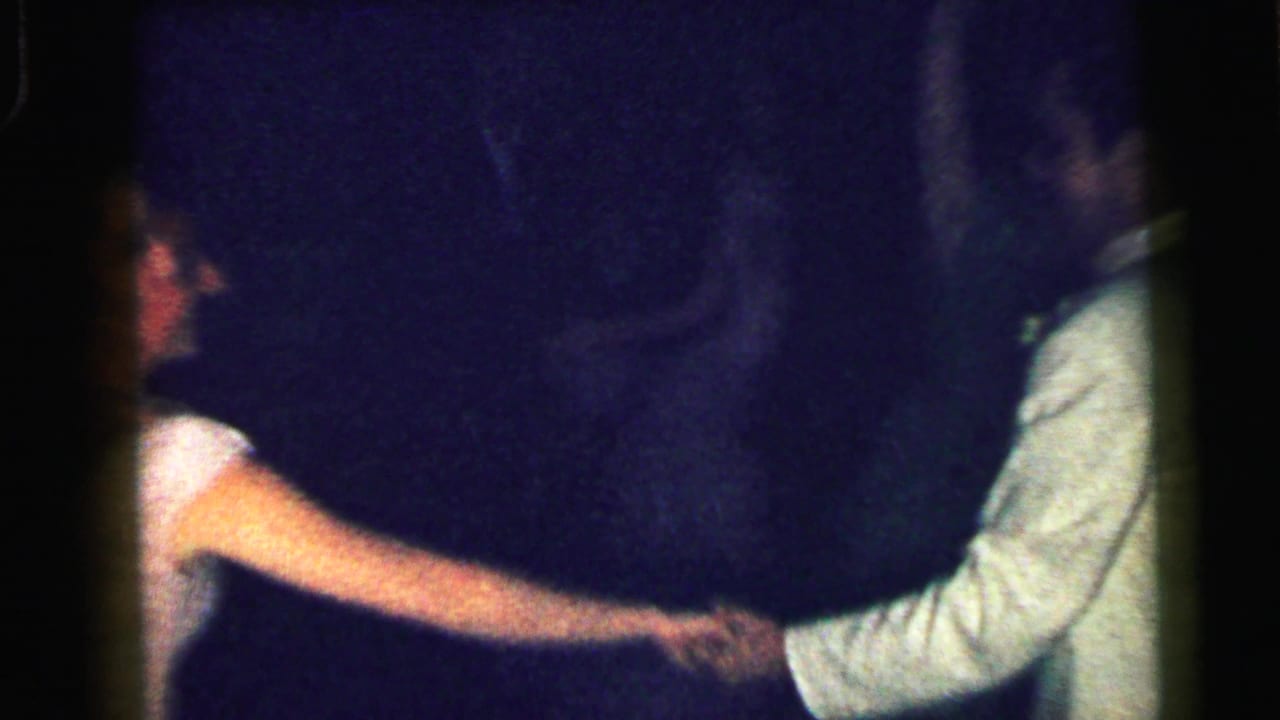 A woman in a pale dress reaches toward a man in a light jacket in a dark dance room.