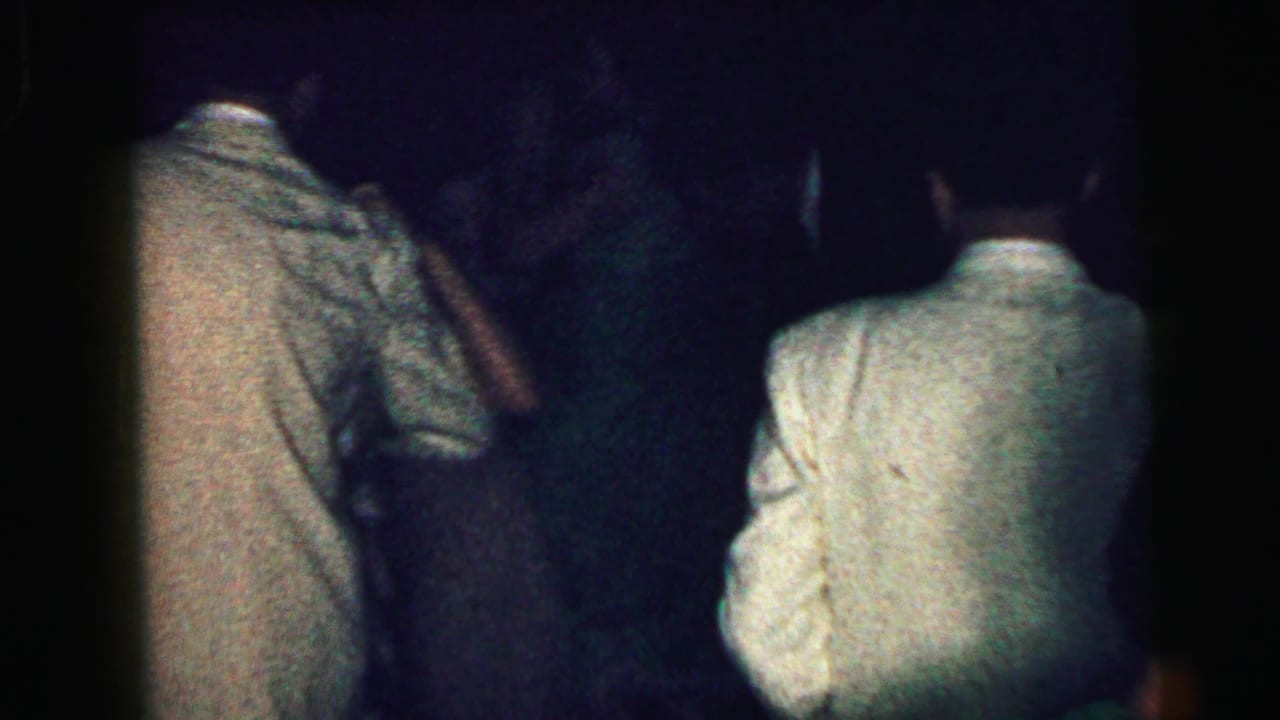Several adults in light jackets and dark suits stand or dance in a dim room.