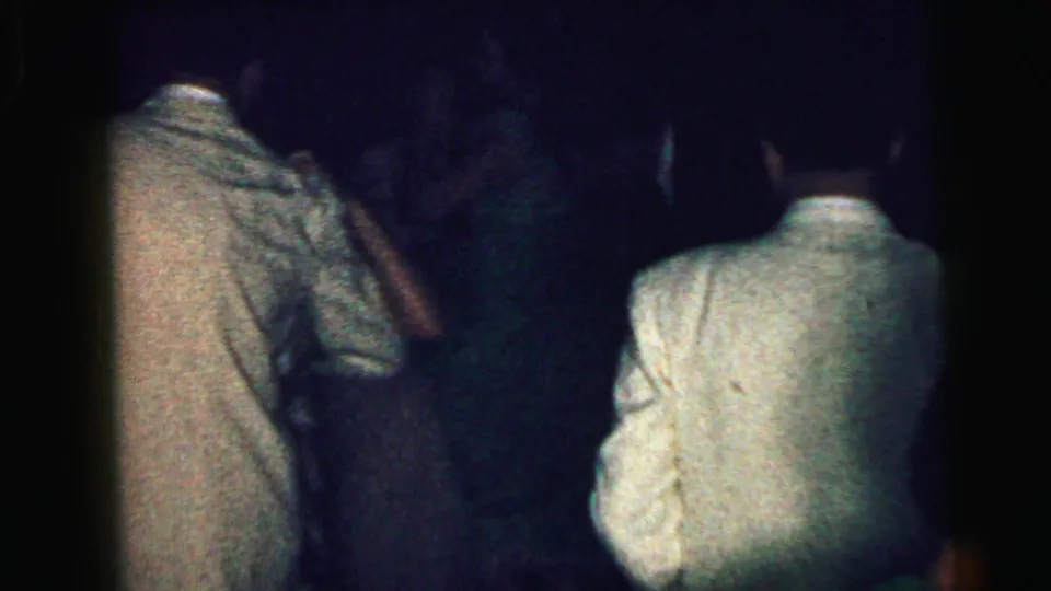 Adults in formal clothing move through a dark Camden 1964 dance setting, with backs and shoulders filling most of the frame.