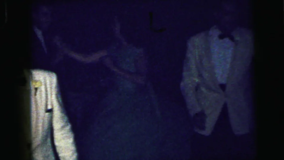 A low-light 8mm frame shows formal-dressed guests on the dance floor, with a white tuxedo jacket partly visible near the center.