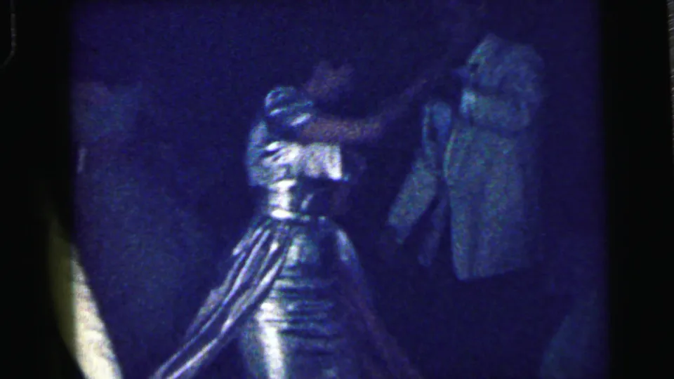 The frame shows a woman in a shiny patterned dress dancing near another formally dressed guest in dark blue 8mm party lighting.