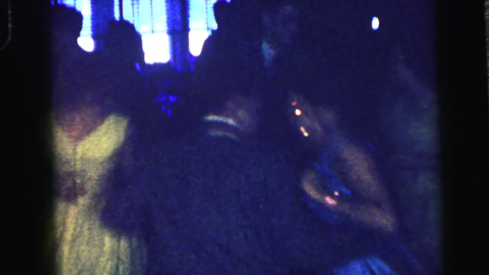 A dark, grainy Camden dance-party frame shows bright lights above a crowded room with guests and partial figures in formal clothing.