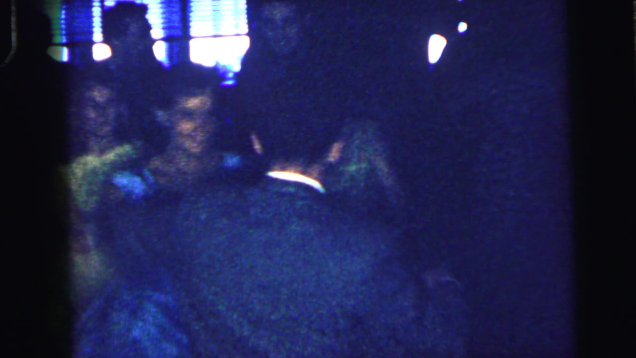 Dimly lit adults standing or dancing in a crowded room under bright window light.