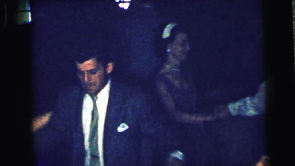 1964 Camden home-movie frame showing a man in a dark suit and tie among dancing guests in a low-lit indoor gathering.