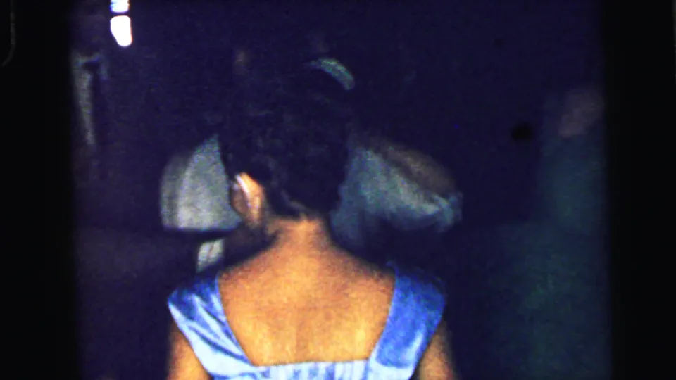 1964 Camden party frame showing the back of a woman in a shiny blue dress facing other guests in a crowded, low-lit room.