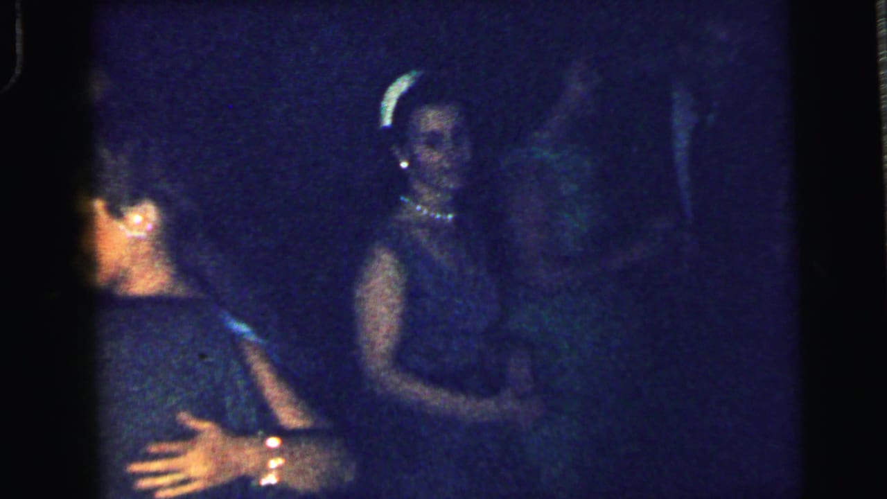 Two formally dressed women appear in conversation or dance under dim party lighting in Camden, New Jersey.