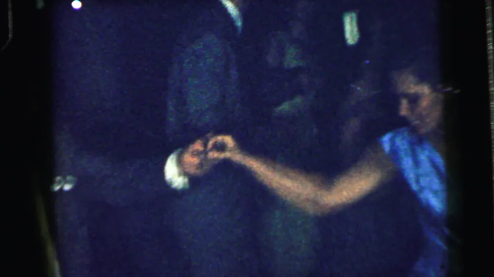 In a 1964 Camden, New Jersey home-movie frame, a woman in a blue dress reaches toward another dancer under low party lighting.