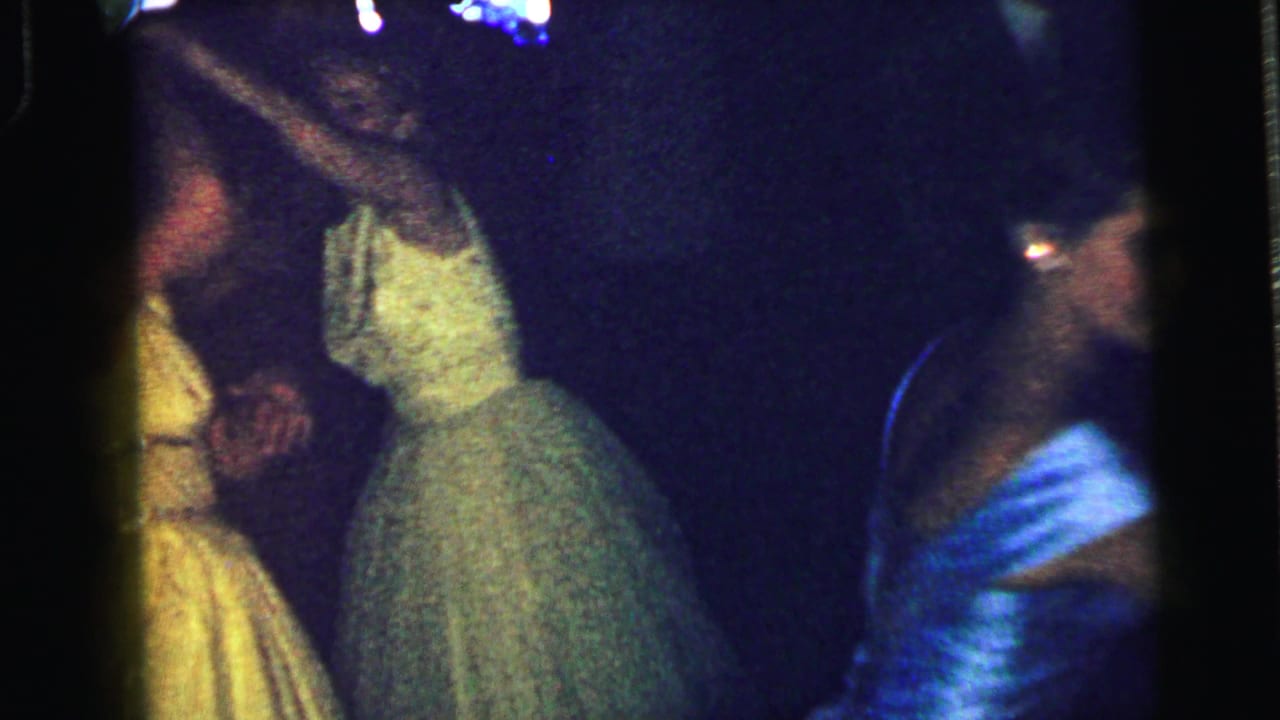 Women in yellow, pale green, and blue dresses dance at the Camden formal party, with a suited man visible in the background.
