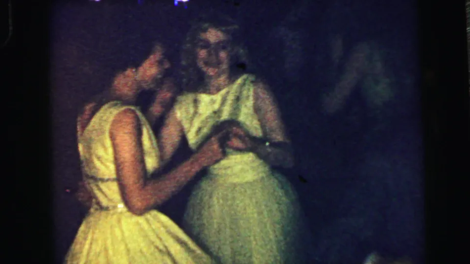 Two women in full-skirted formal dresses dance closely in the dim Camden party footage, one in yellow and one in pale green.