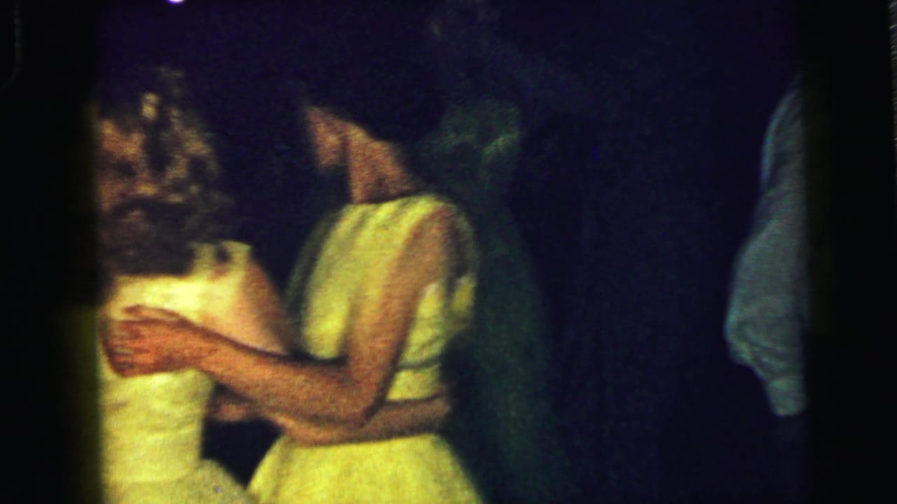 Couple dancing closely in dim light, with a woman in a bright yellow dress near the center.