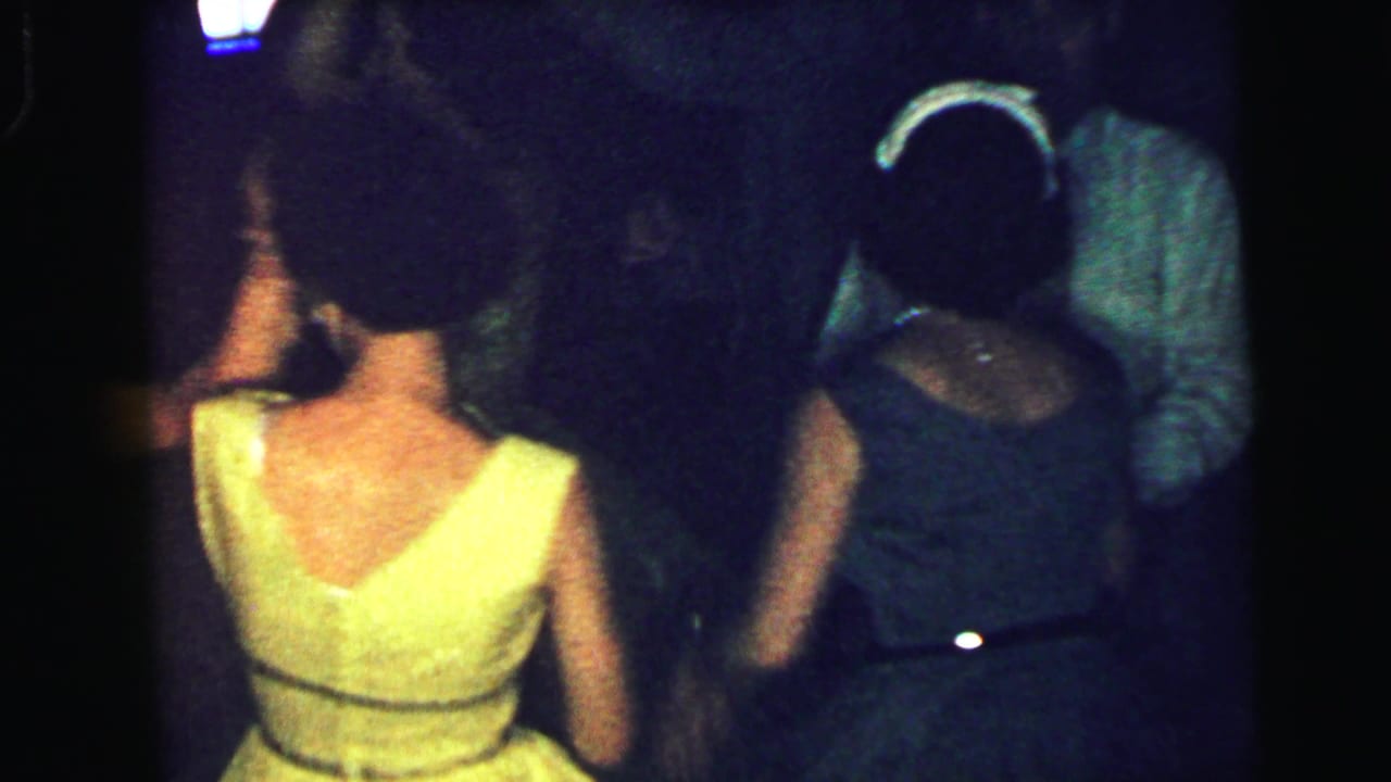 Woman in a yellow dress seen from behind while other adults dance in a dark room.