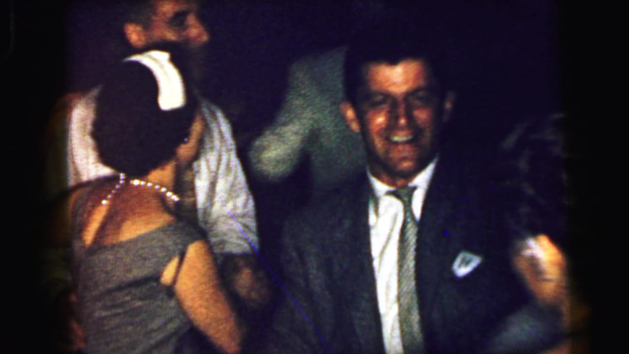 Wedding guests dancing in a dim reception room, with a smiling man in a suit near the center.