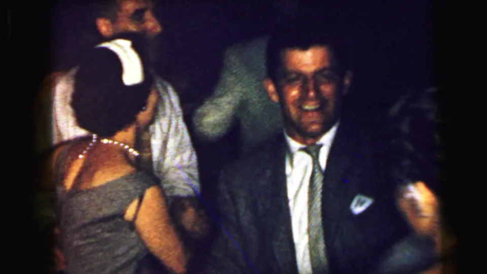 1964 Camden home-movie frame showing wedding guests dancing indoors, including a smiling suited man and partially visible women around him.