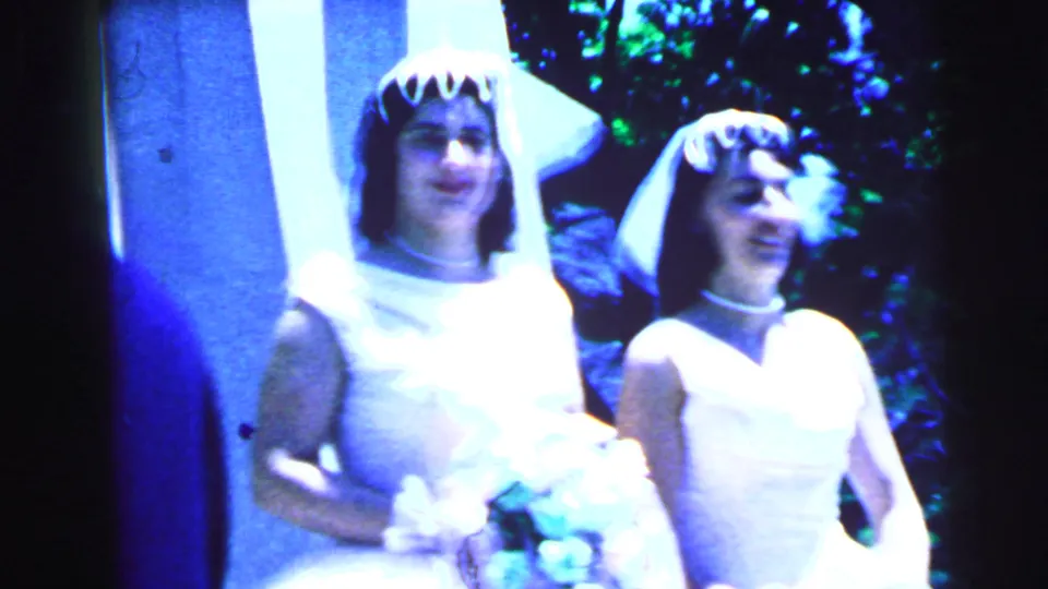 1964 Camden frame showing two veiled women in white wedding dresses posing outdoors, one holding a light-colored flower bouquet.