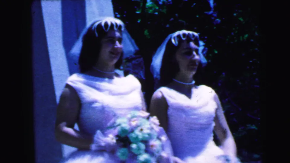 Two veiled women in white wedding dresses stand together outdoors in 1964 Camden footage, with a bouquet visible in the foreground.