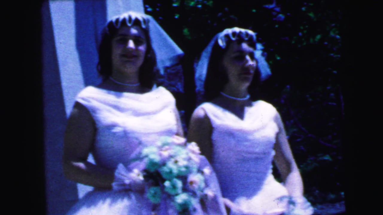 Two brides in white dresses and veils stand with bouquets near a building entrance.