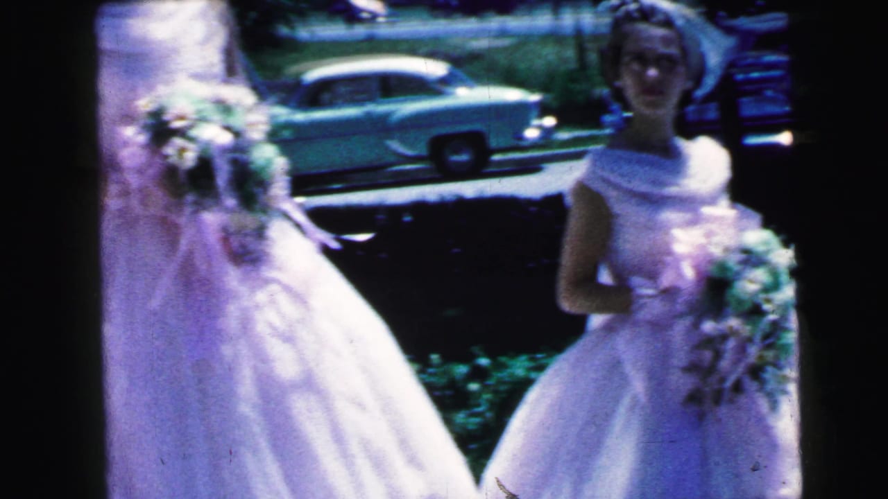 Young women in white formal dresses hold bouquets outdoors near parked cars.