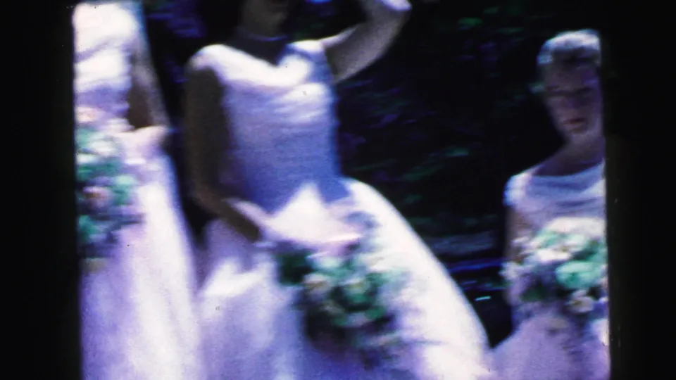Soft 1964 home-movie frame of young women in white formal dresses holding bouquets, with one face partly visible near the right side.