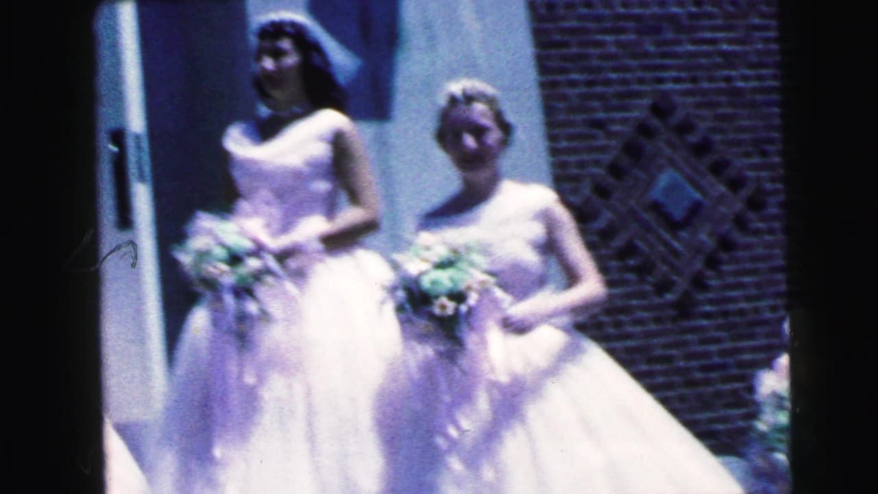 Two bridesmaids in white formal gowns stand with floral bouquets outside a brick church in Camden, New Jersey, 1964.