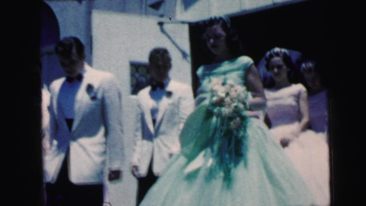 1964 Camden home-movie frame showing a wedding party outside, including a bride in a green gown, bouquet, white tuxedo jackets, and formal dresses.