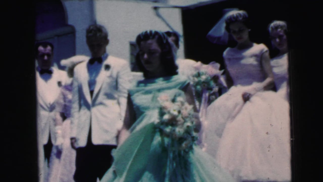 Bride in green gown walks with groom and attendants, with bridesmaids in pale formal dresses behind them.