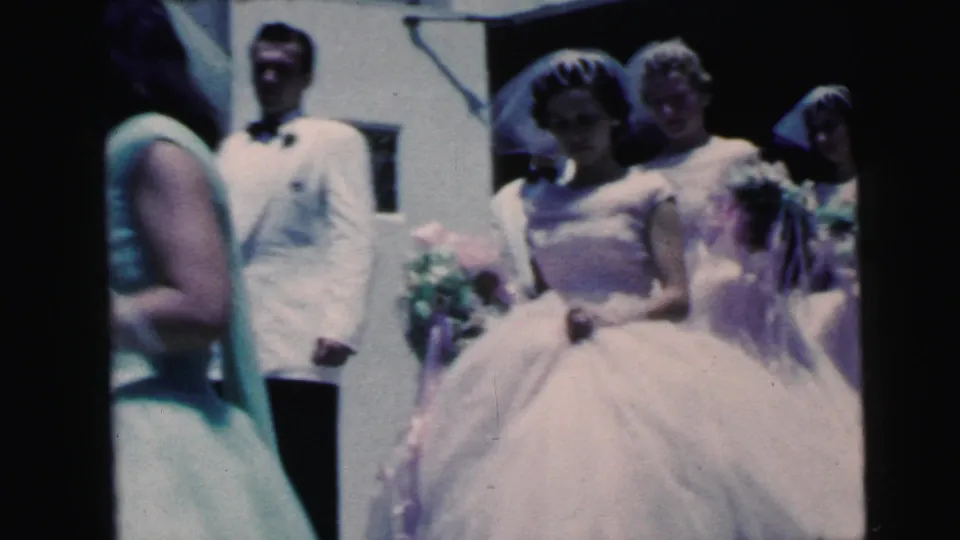 1964 Camden home-movie frame showing a bridesmaid in a full pale dress and veil near tuxedoed attendants as the bride crosses the foreground.