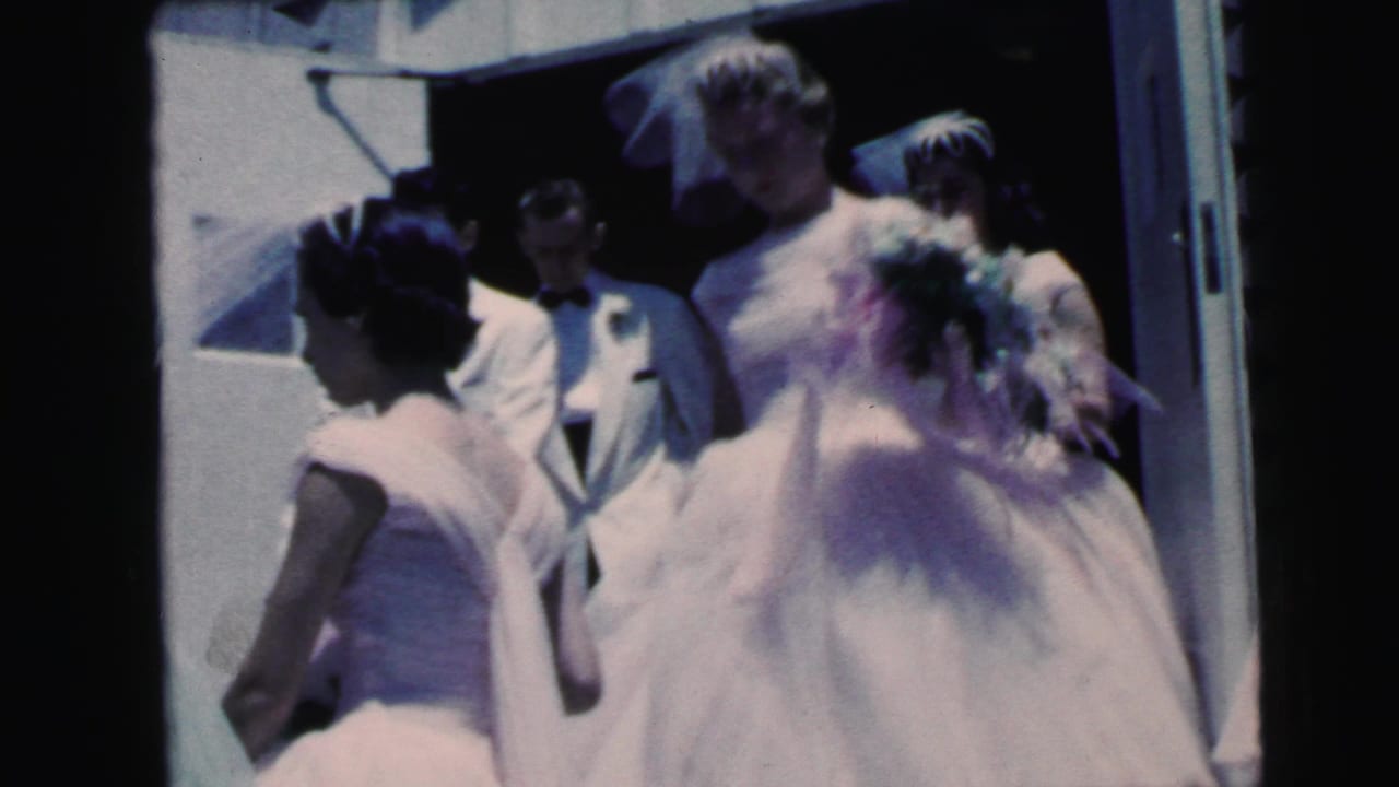Wedding party members gather outside a doorway, with a bride in a veil seen from behind.