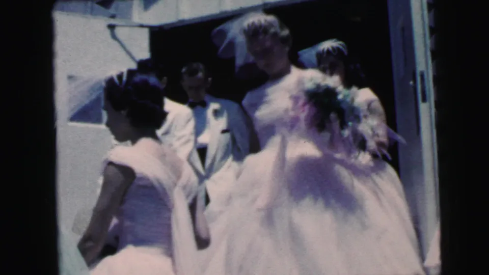 1964 Camden wedding home-movie frame showing the bride from behind near bridesmaids and groomsmen at a bright exterior entrance.