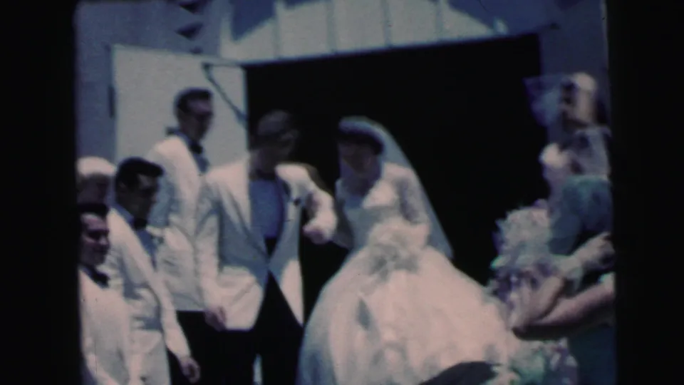 1964 Camden wedding home-movie frame showing a bride and groom leaving a church doorway among formally dressed guests.