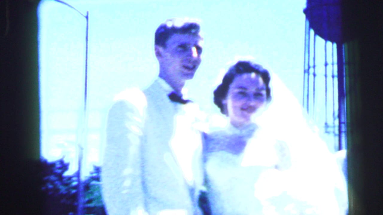 1964 Camden wedding home-movie frame showing a bride and groom posing outdoors in strong blue light, with veil and bouquet visible.