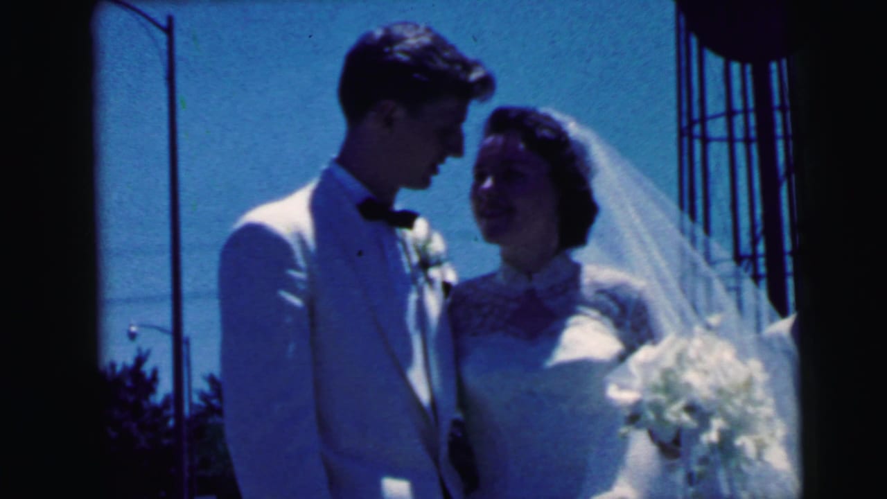 Bride and groom in 1964 Camden footage look toward each other under a blue sky, with the bride holding flowers and wearing a veil.