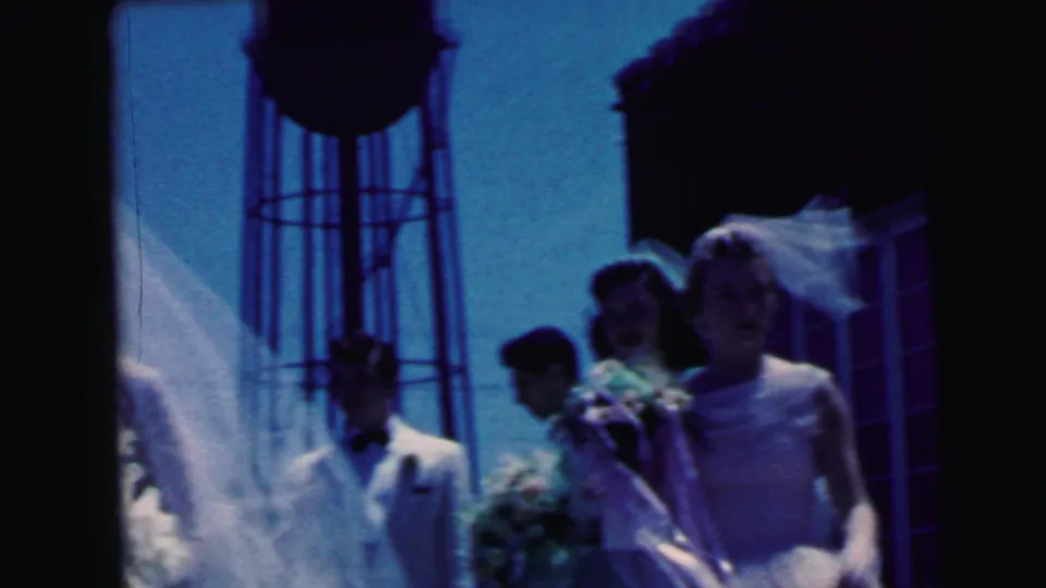 Bride in 1964 Camden wedding footage stands with bouquet and veil while other wedding party figures gather beside her.