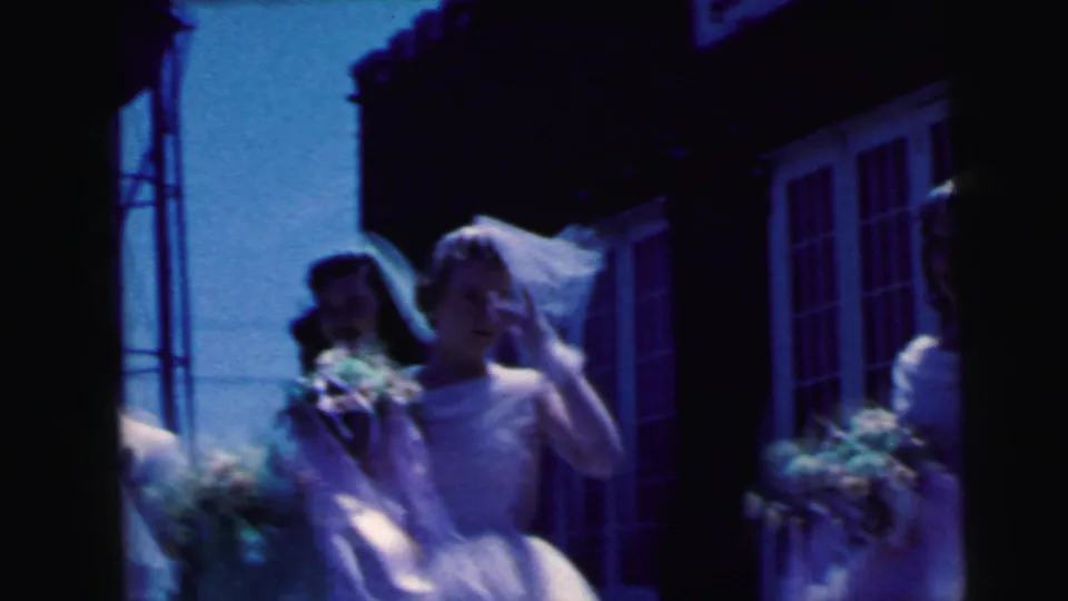 1964 Camden home-movie frame showing bridesmaids in white dresses and veils waiting near a brick venue with bouquets.