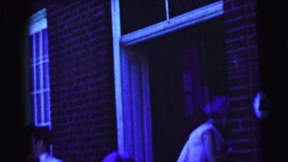 1964 Camden home-movie frame showing a veiled woman in a white dress beside a brick venue doorway and window.