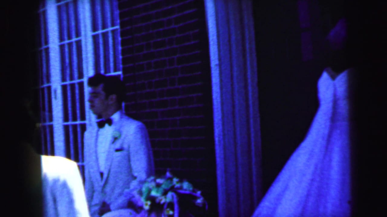 1964 Camden wedding footage shows a groom in a light suit and bow tie beside a bride in a white gown near the church entrance.