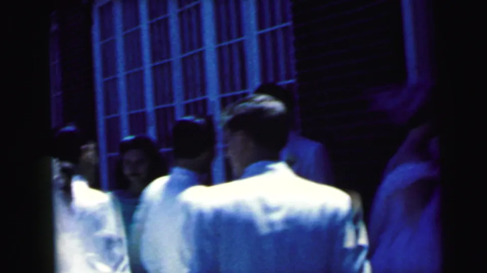 Wedding guests and attendants move through a narrow exterior space near tall windows during the 1964 Camden church arrival.