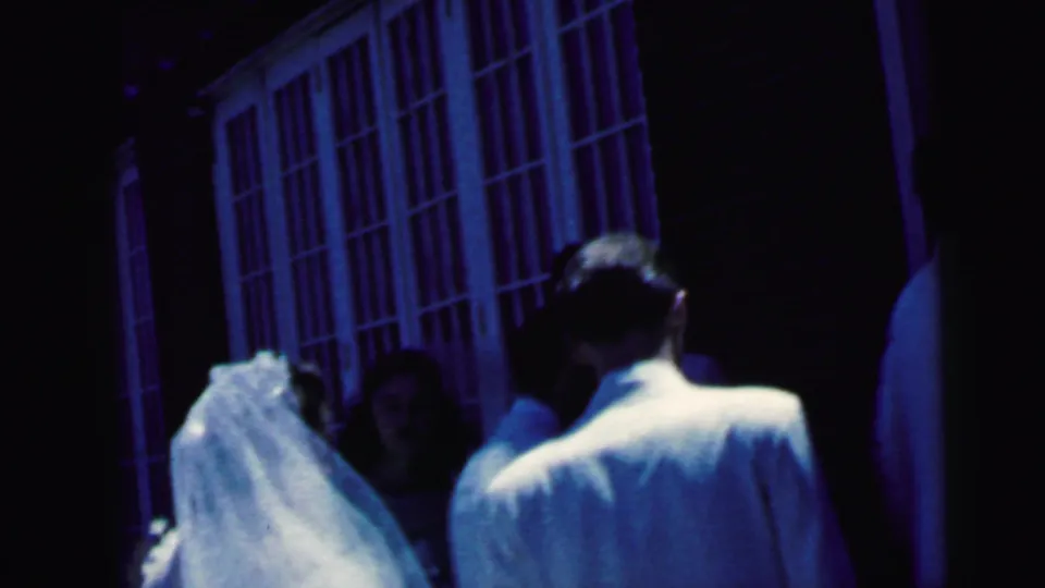 The veiled bride and a man in a light suit stand near the church doorway in a dim 1964 Camden wedding home movie frame.
