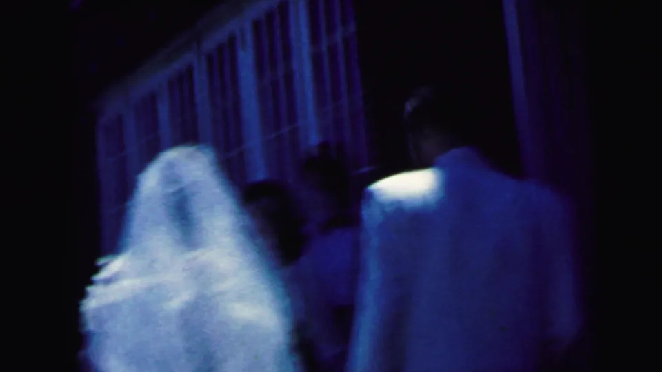 1964 Camden home-movie frame showing a bride in a white veil and groom in a light jacket approaching a dark church entrance.