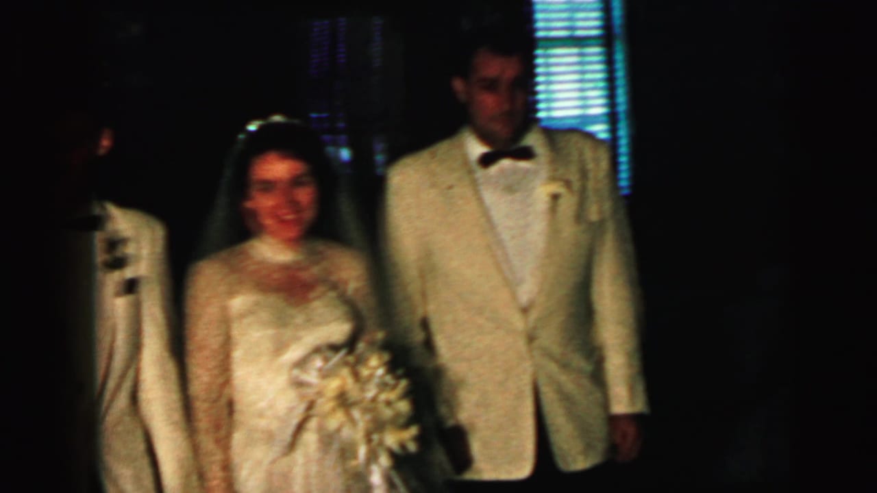 Bride holding a bouquet stands beside groom in a cream tuxedo in a dim interior.