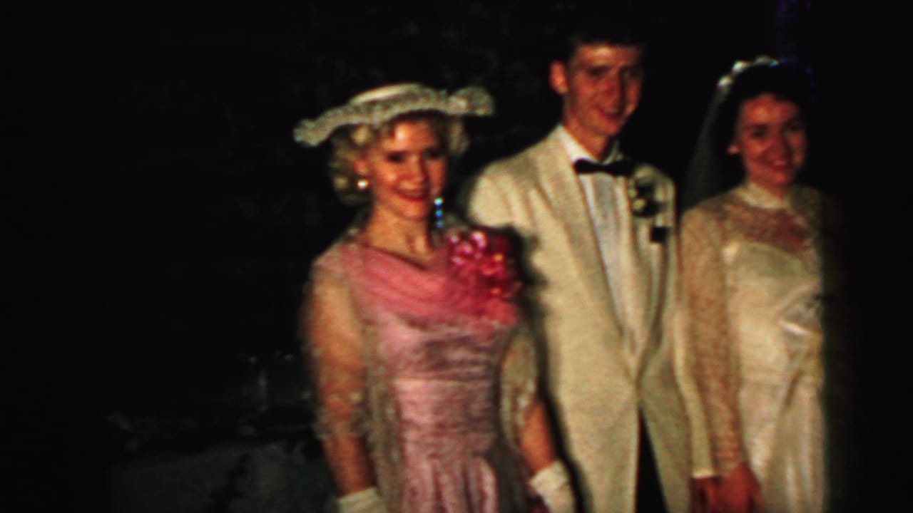 1964 Camden wedding frame with a pink-dressed guest, tuxedoed man, and bride smiling in a dark interior.